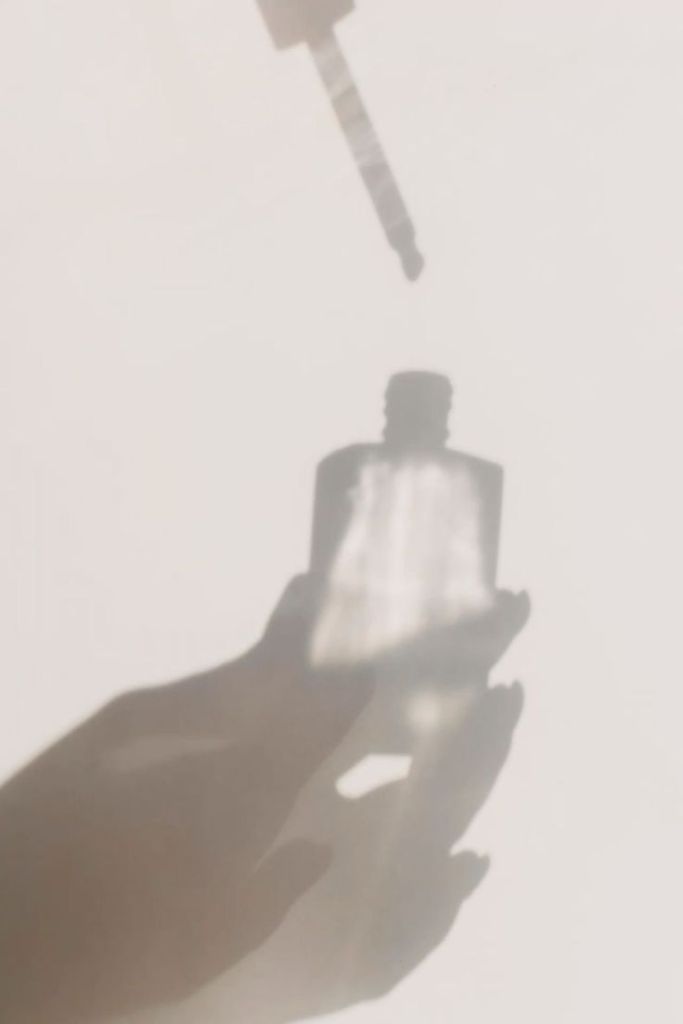 Shadow of a hand holding a bottle with a dropper above it, casting a soft silhouette on a light background.