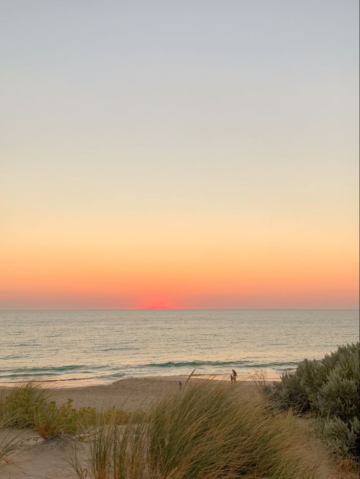 A serene beach view at sunset, with gentle waves lapping at the shore and a gradient sky transitioning from light blue to soft pink and orange, framed by grass and shrubs.